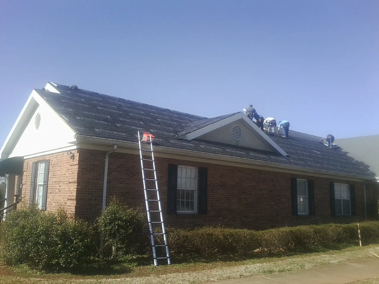 Crew tearing shingles off a brick ranch roof with a ladder set — Emergency Roof Repair work by our crew in Pell City