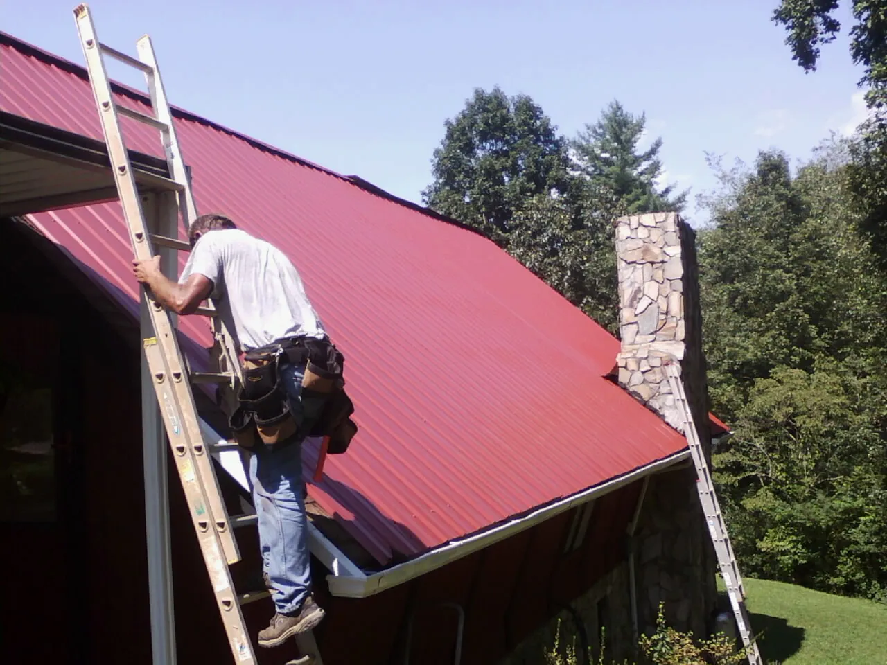Roofer on a ladder beside a red metal roof and stone chimney — Soffit & Fascia Repair by Roofing Brothers in Pell City